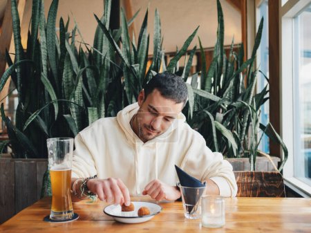 Un joven en una pose relajada disfruta de sus aperitivos y un vaso de cerveza, sentado cómodamente en un café rodeado de vibrantes plantas de interior