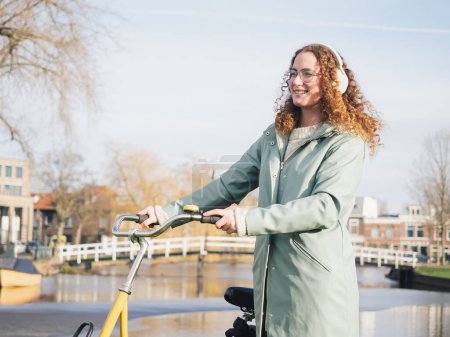 Eine fröhliche junge Frau mit lockigem Haar und Kopfhörern auf einem gelben Fahrrad neben einem malerischen Kanal in einer sonnigen urbanen Umgebung