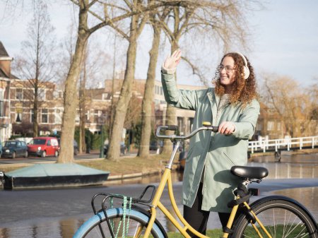 Ein Bild zeigt eine fröhliche Frau mit lockigem Haar und Kopfhörer, die Freunden zuwinkt, während sie neben ihrem gelben Fahrrad in der Nähe eines malerischen Kanals steht.