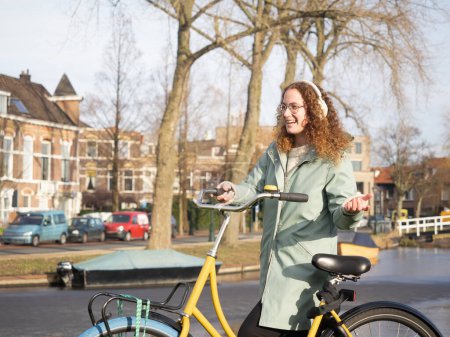 Zu sehen ist eine überschwängliche junge Frau mit lockigem Haar und Brille, die neben ihrem gelben Fahrrad spaziert und in einer malerischen Straße mit historischen Amsterdamer Häusern im Hintergrund ihrer Freude Ausdruck verleiht.