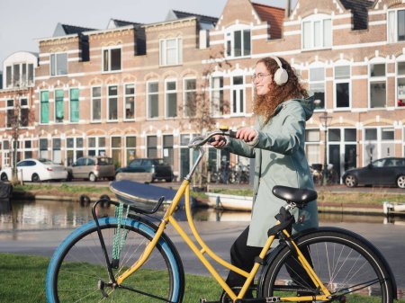 Eine fröhliche Frau mit lockigem Haar und Kopfhörern genießt einen sonnigen Tag, als sie mit ihrem gelben Fahrrad an einem Kanal in Amsterdam spaziert, im Hintergrund klassische holländische Häuser