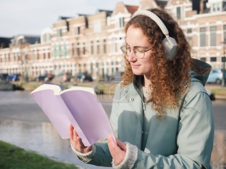 Eine fokussierte junge Frau mit lockigem Haar und Brille, die Kopfhörer trägt, liest ein Buch am Kanal, im Hintergrund die klassische niederländische Architektur.