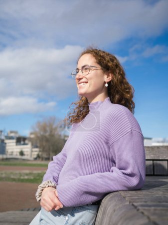 Captured in a gentle moment, a woman adorned in a lavender sweater radiates contentment, her gaze fixed on the distant horizon, against a vivid sky and urban backdrop