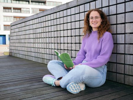 A cheerful curly-haired woman in glasses sits cross-legged, enjoying a book outdoors with buildings in the background looking at camera