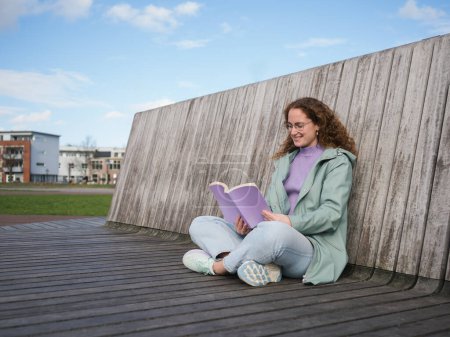 Une femme aux cheveux bouclés et aux lunettes s'assoit les jambes croisées en lisant un livre, avec un mur en bois rustique et un paysage urbain derrière elle