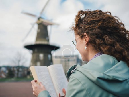 Blick über die Schulter einer Frau mit lockigem Haar, die ein Buch liest, mit einer traditionellen Windmühle im verschwommenen Hintergrund