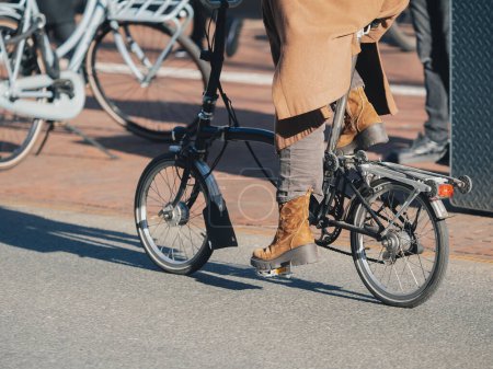 Urban scene with an individual riding a foldable bike, featuring stylish brown boots and coat in Amsterdam
