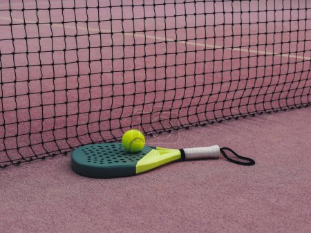 Photo for A yellow and black padel racket with one yellow ball resting on a clay-colored court beside a black net - Royalty Free Image