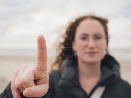A sharp focus on a single finger gesturing 'one' with a blurred figure and beach in the background