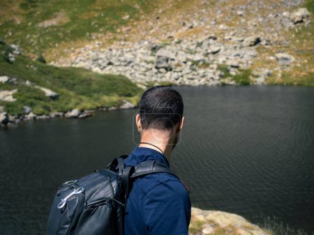 Un caminante masculino con una mochila mira a un lago tranquilo rodeado de terreno accidentado en un entorno de montaña