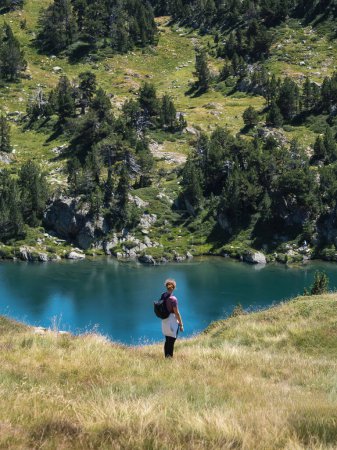 A woman with a backpack stands on a grassy hill, admiring the view of a tranquil blue lake surrounded by green forest. Pyrenees