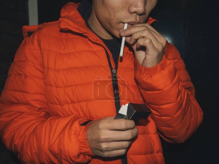 Close-up image capturing a man in an orange puffer jacket as he lights a cigarette, highlighting the action of smoking