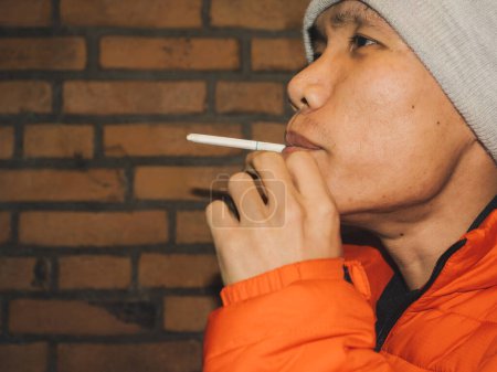 Nahaufnahme eines nachdenklichen jungen asiatischen Mannes, der eine Zigarette raucht, mit einer grauen Mütze und orangefarbener Jacke vor einer Backsteinmauer steht