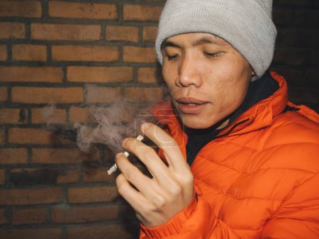 Nahaufnahme eines jungen asiatischen Mannes, der Rauch einer Zigarette ausatmet, bekleidet mit orangefarbener Jacke und grauer Mütze, vor einer rustikalen Ziegelwand