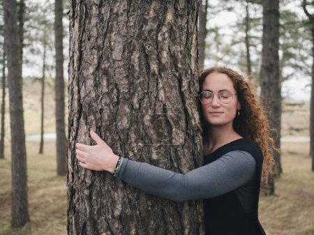 Middle-aged woman with curly hair and glasses peacefully embracing a tree in a calm forest environment