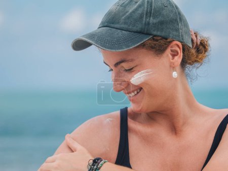 Joyful woman applying sunscreen on her cheek, demonstrating skin care on a bright beach day