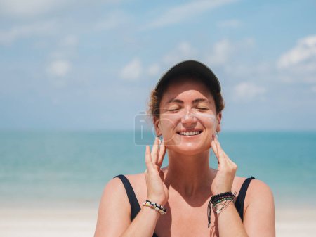 Happy woman in a black swimsuit and cap applying sunscreen on her face while standing at the beach
