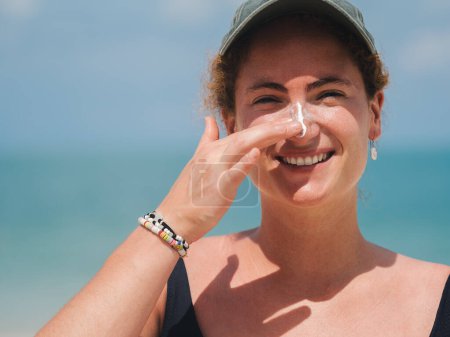 Smiling woman in a black swimsuit and cap applying sunscreen to her nose while enjoying a sunny day at the beach. Concept of skin protection