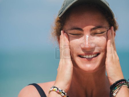 Smiling woman in a black swimsuit and cap applying sunscreen to her face while enjoying a sunny day at the beach
