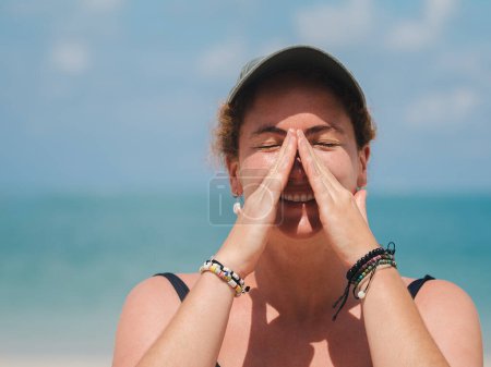 Close-up of a woman smiling with her eyes closed, applying sunscreen to her nose at the beach. She wears a hat and bracelets, enjoying a sunny day by the ocean