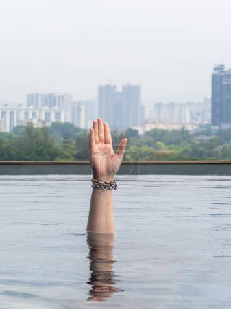 A hand wearing bracelets is raised above the water in an infinity pool, suggesting a call for help, with a city skyline in the background