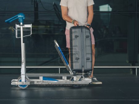Man in casual clothes and pink shorts placing a large suitcase onto an airport luggage trolley outside a terminal with reflective glass windows. Travel preparation concept