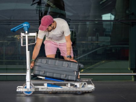 Casually dressed man in a red cap adjusts a large black suitcase on an airport luggage trolley outside a terminal with reflective glass windows. Travel preparation scene