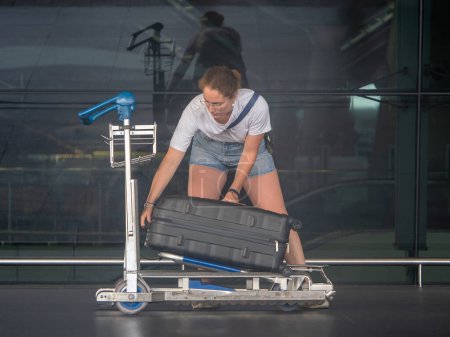 Woman in casual attire and glasses loads a large black suitcase onto an airport luggage trolley outside a terminal with reflective glass windows. Travel preparation scene