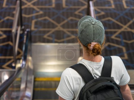 Vue arrière d'une femme avec un sac à dos et une casquette montant un escalier roulant dans un terminal de l'aéroport. Voyages et transports