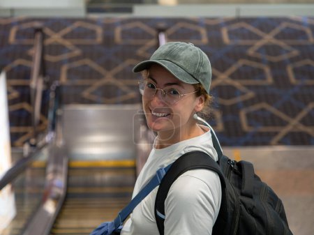 Smiling woman wearing glasses, a cap, and a backpack on an escalator inside an airport terminal. Travel and transportation theme