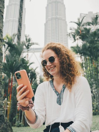 Smiling woman with curly hair and sunglasses using her smartphone for a video call in an urban park with petronas towers in the background. Kuala Lumpur, Malaysia