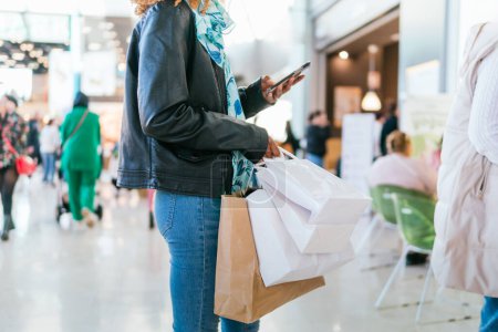 Unrecognizable black girl looks at her phone while queuing in a shopping center, carrying shopping bags and many people are seen out of focus in the background of the image. No face is visible