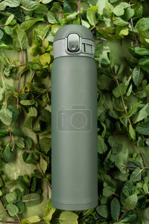 Téléchargez les photos : Thermos mug on a green background. Thermos and mint leaves. Vertical frame. - en image libre de droit