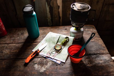Photo for Compass, knives and a map on the table. Thermos and mug on the table. Tourist in a hut by the window. View from above. - Royalty Free Image