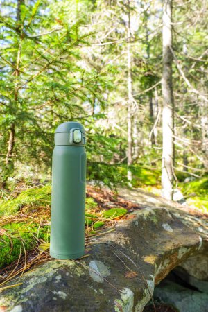 Photo for Green thermos on the stone. Thermal bottle on the background of the forest. Sunny day in the forest. Vertical frame. - Royalty Free Image