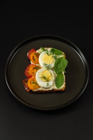 Téléchargez les photos : Sandwich aux œufs durs et tomates. Sandwich dans une assiette. Plaque noire et fond sombre. Cadre vertical. - en image libre de droit