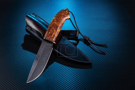 Téléchargez les photos : Couteau de chasse en acier Damas. Couteau Premium avec gaine en cuir. Couteau et lanière en cuir. Fond en carbone avec rétro-éclairage bleu.. Vue avant oblique. - en image libre de droit