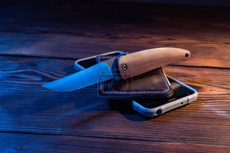 Ein scharfes Klappmesser liegt auf einer Brieftasche und einem Smartphone. Ein scharfes Messer mit Holzgriff. Blick von oben schräg.