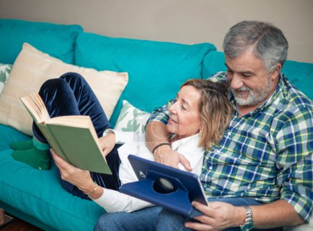 An affectionate retired couple is captured relaxing on the sofa in their cozy living room, engrossed in reading a book and watching movies on a tablet. This heartwarming image depicts the simple joys of retirement