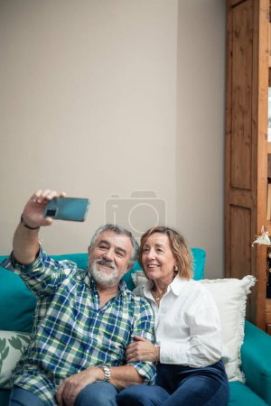 In einem herzerwärmenden Moment zu Hause sitzt ein Rentnerpaar gemütlich auf seinem Sofa und hält mit seinem Handy ein Selfie fest. Mit einem Lächeln im Gesicht umarmen sie die Freude der Gegenwart, 