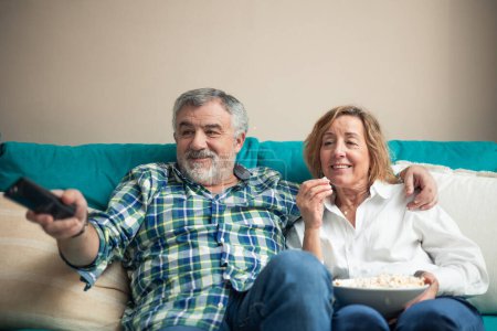 In the comfort of their home, a senior couple sits together on the sofa, immersed in a movie on television. With a bowl of popcorn in hand and the TV remote pointed at the screen, they share a delightful evening, 