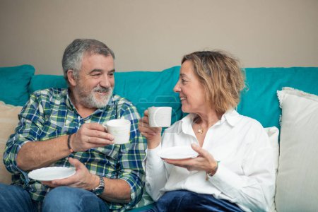 Basking in the warmth of their home, a senior couple sits cozily on the sofa, savoring cups of coffee with radiant smiles. Their joy is palpable as they share tender moments of companionship and relaxation. 