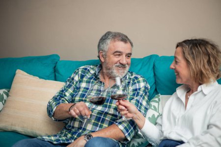 Amidst the cozy ambiance of their living room, a senior couple shares a heartwarming toast with glasses of wine on the sofa. Their smiles reflect the depth of their love and the joy of shared moments. 