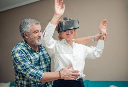 Immersed in the wonders of modern technology, a senior couple enjoys a playful moment in their living room. The woman wears virtual reality glasses, transported to a digital realm, while her husband gently supports her from behind