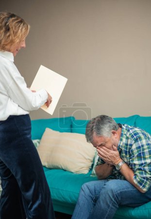 In a poignant scene within their home, a senior couple grapples with turmoil as the woman gestures angrily towards documents, her expression marked by frustration, while the husband, seated on the sofa, covers his face with his hands