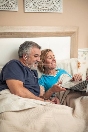 Ein älteres Ehepaar entspannt es sich bequem zu Hause, liegt mit Kopfhörern im Bett und einem Laptop auf dem Schoß. Das Bild spiegelt die Integration der Technologie in den Alltag älterer Erwachsener wider 