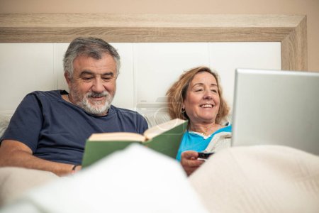 In dieser charmanten Szene genießt ein älteres Paar den gemeinsamen Abend im Bett. Der eine ist in die digitale Welt vertieft und arbeitet am Laptop, während der andere Trost in den Seiten eines fesselnden Buches findet.. 