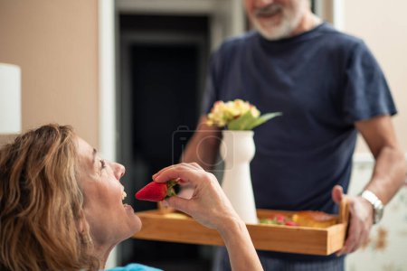 This captivating image brings us up close to a serene moment in the morning routine of a elderly retired couple. The focus is on the woman, who is delightfully indulging in a fresh strawberry while reclining comfortably in bed. 