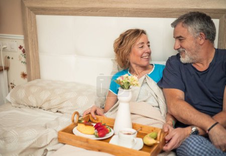 Dieses schöne Bild fängt einen intimen Moment zwischen einem älteren Paar ein, das im Bett frühstückt. Mit einem Blick voller Liebe und Intimität erfreuen sie sich an der Gesellschaft des jeweils anderen, während sie diesen besonderen Moment teilen.. 