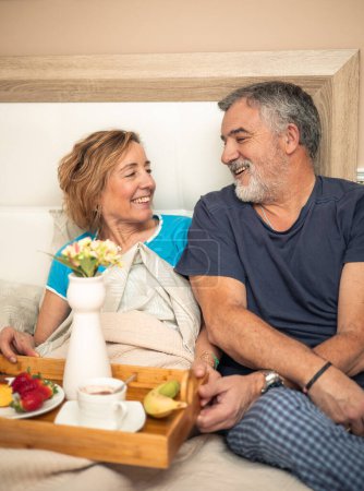 Dieses herzerwärmende Bild fängt die liebevolle Verbundenheit zwischen einem älteren Paar ein, das sich das Frühstück im Bett teilt. Während sie ein Lächeln voller Wärme und Zuneigung austauschen, spiegelt ihr Blick die anhaltende Liebe wider, die sie teilen. 